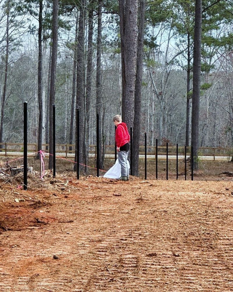 Person in red jacket stands by black fence posts in a wooded area, holding white material.
