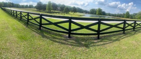 Black wooden fence curving across a grassy field under a cloudy blue sky.