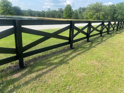 Black wooden fence in a grassy field, with trees in the background under a blue sky.