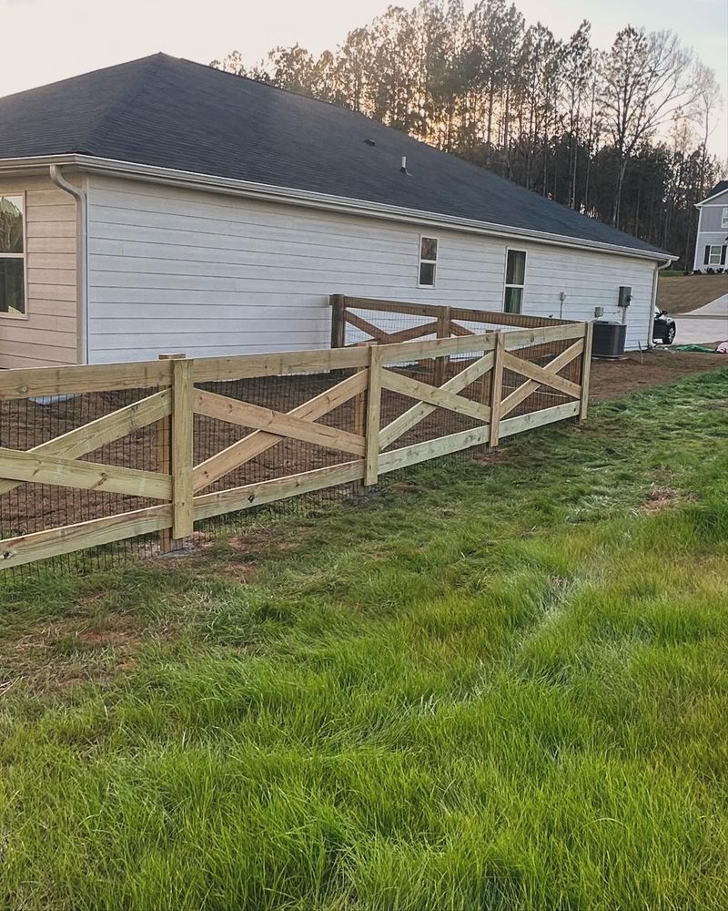 Wooden X-style fence with wire mesh in front of a white house on a grassy lawn.