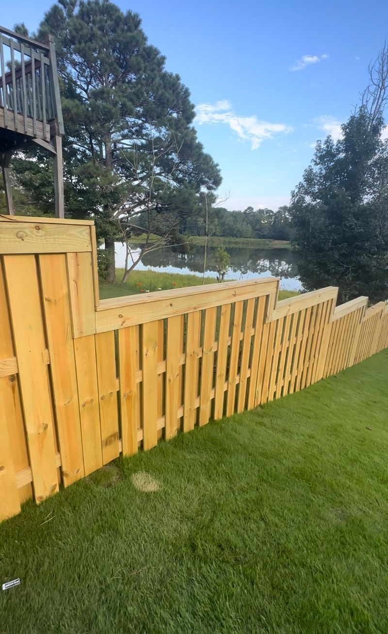 Wooden fence on green grass, leading toward a lake and trees.