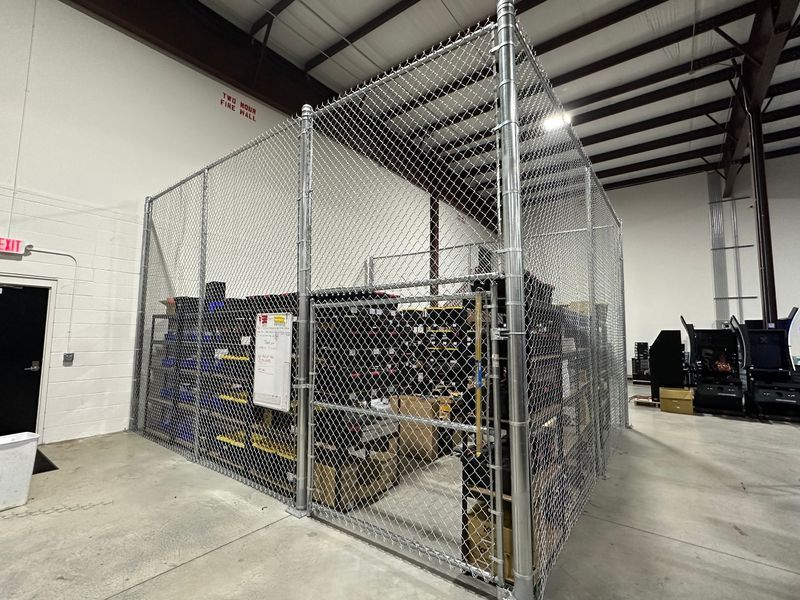 Metal chain-link enclosure within a warehouse. Shelves inside hold boxes and items.