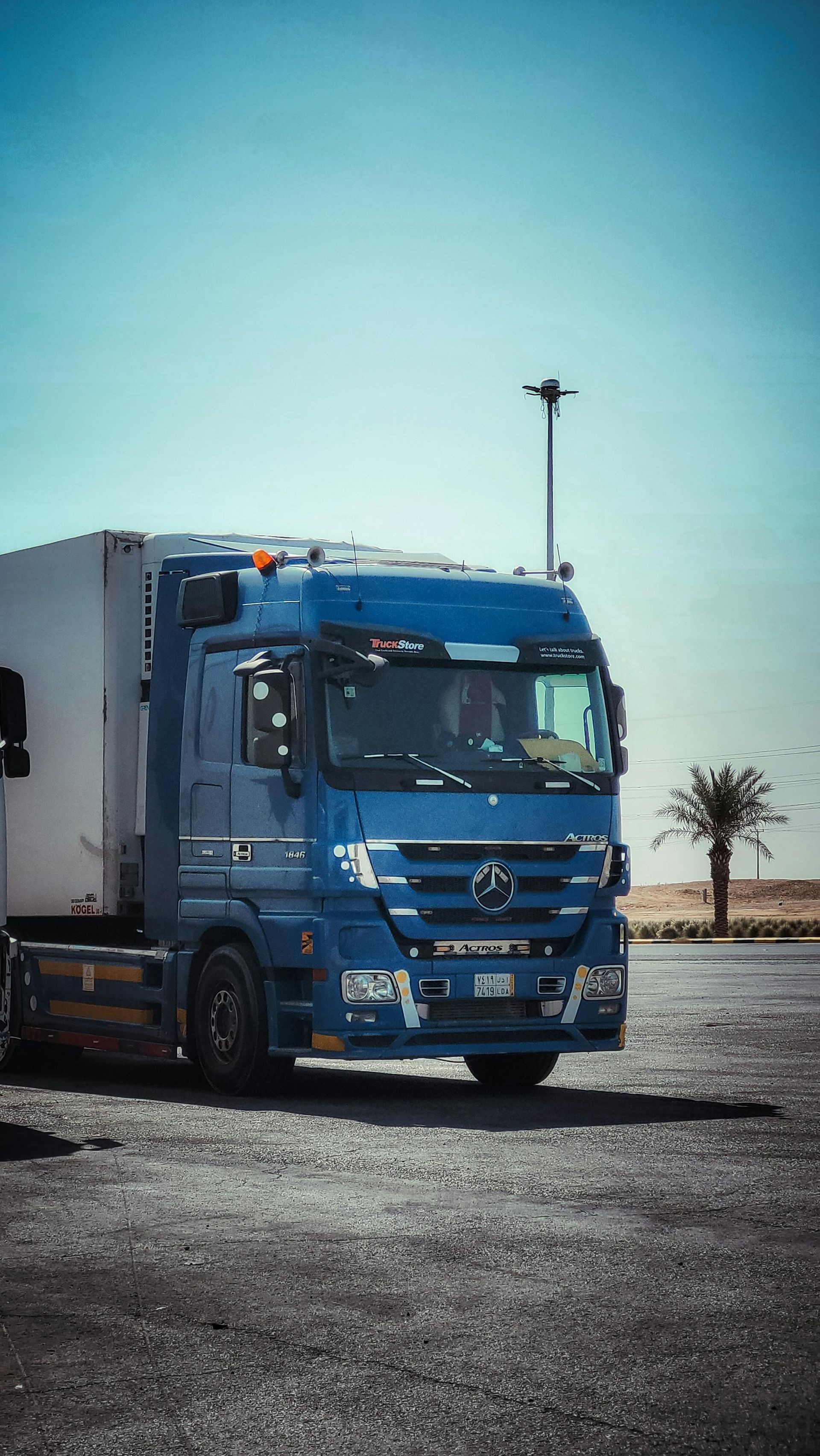 A blue semi-truck with a white trailer is parked on a gravel surface under a clear blue sky.
