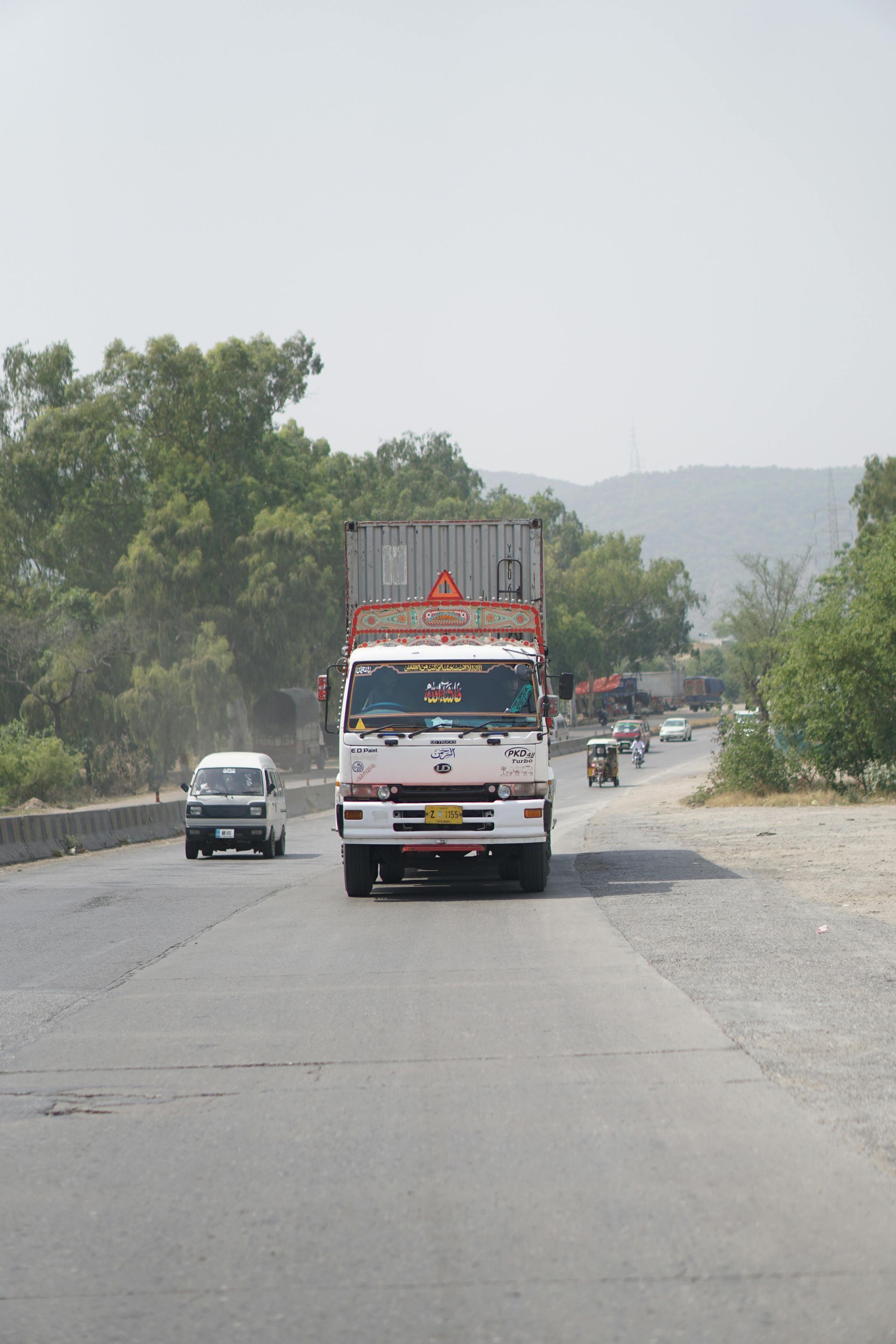A white truck carrying a shipping container drives down a highway lined with trees on a sunny day.