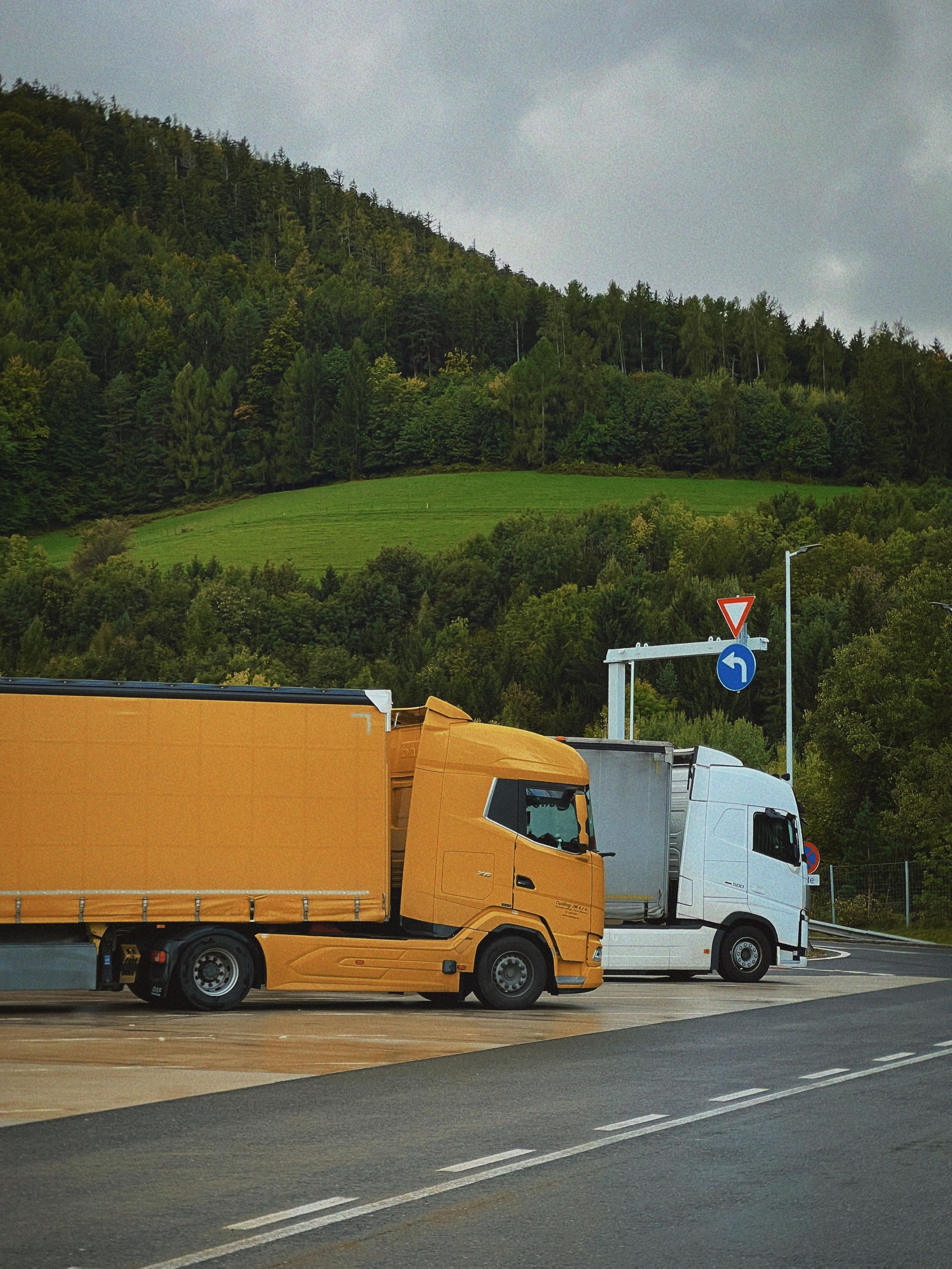 A yellow semi-truck parked behind a white semi-truck on an asphalt road in front of a hillside covered in trees.