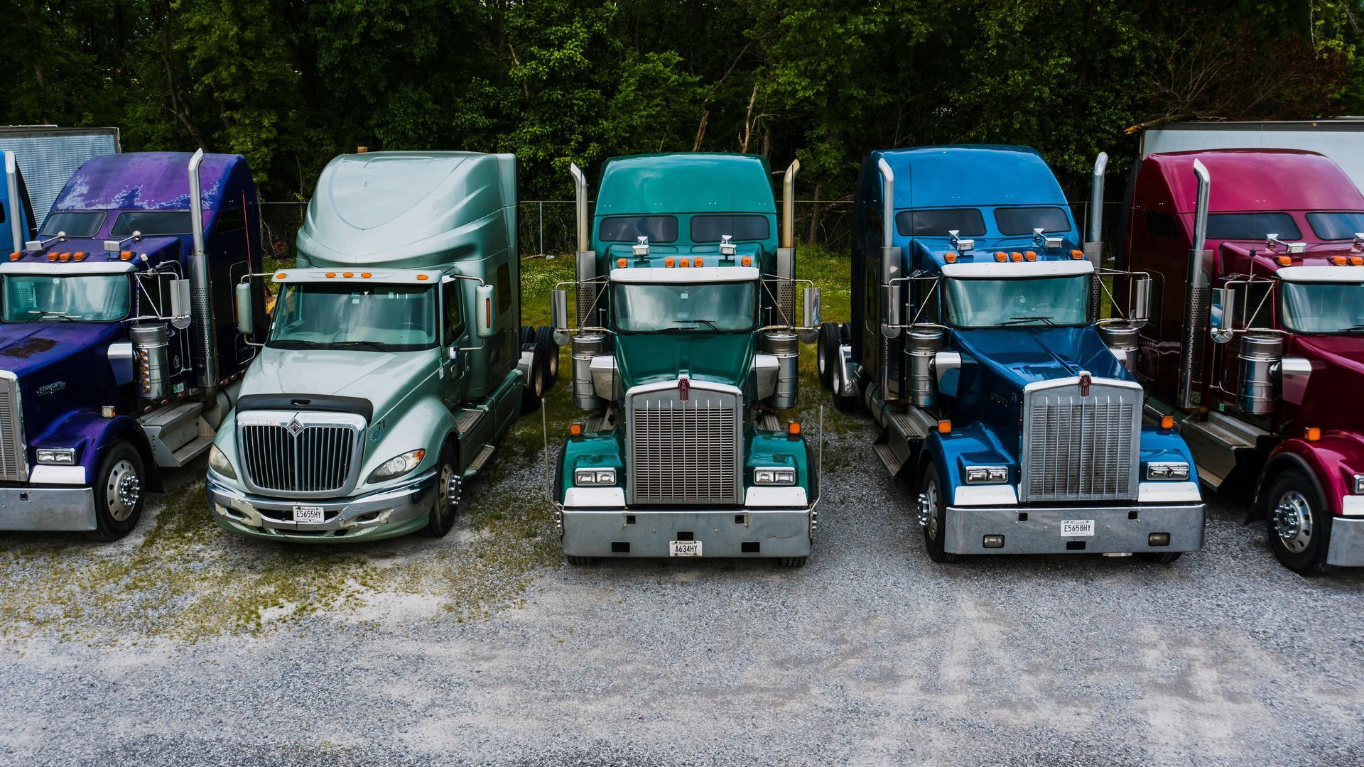 A row of five semi-trucks in purple, silver, green, blue, and maroon parked on a gravel lot in front of trees.