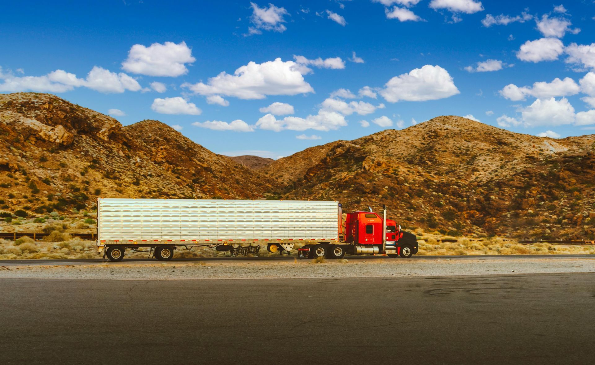 A red semi-truck with a long white trailer drives along a road against a backdrop of desert mountains and a blue sky.