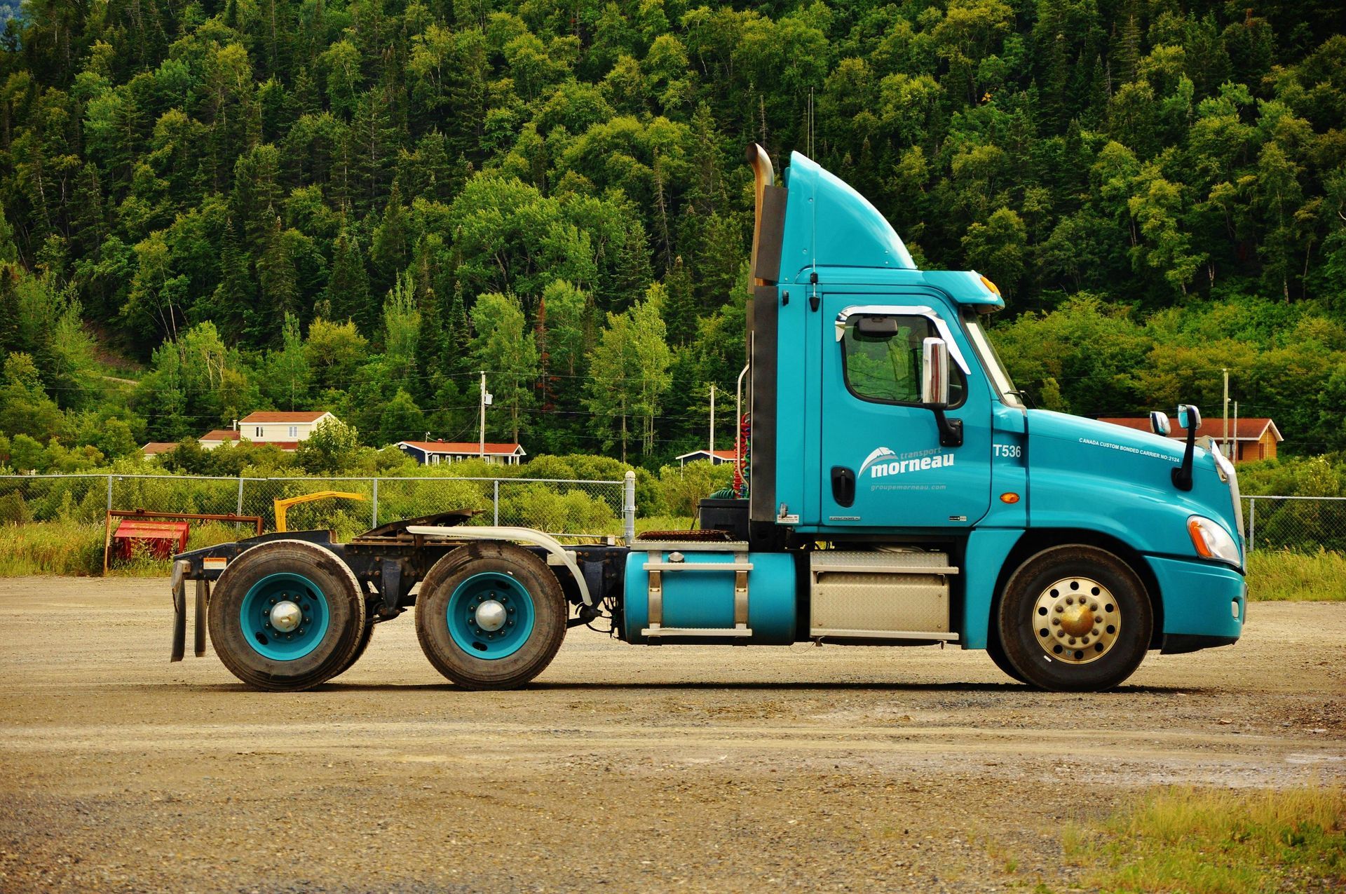A teal semi-truck cab parked on a gravel lot in front of a dense, green forest.