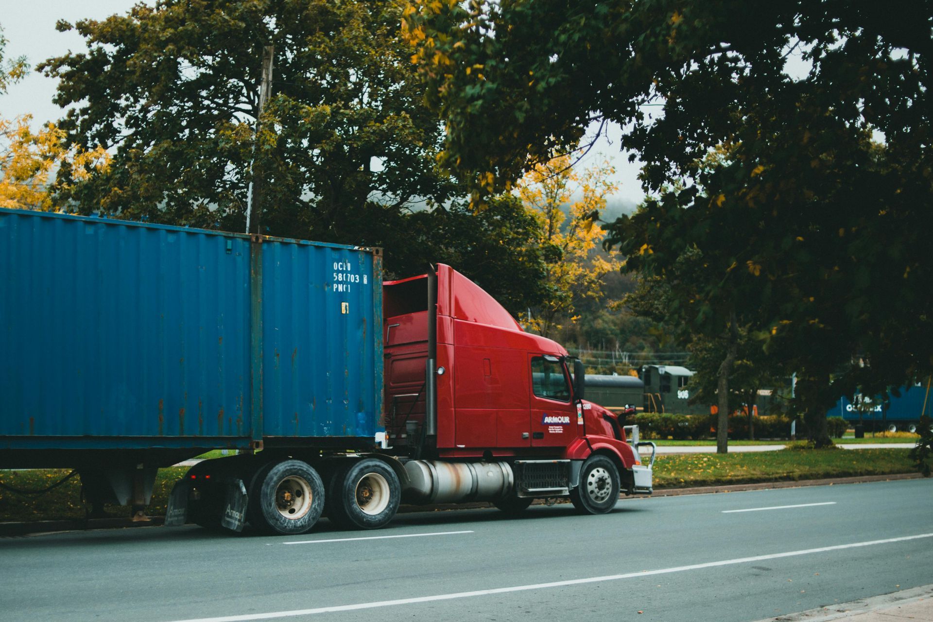 A red semi-truck pulling a blue shipping container driving along a road next to trees.