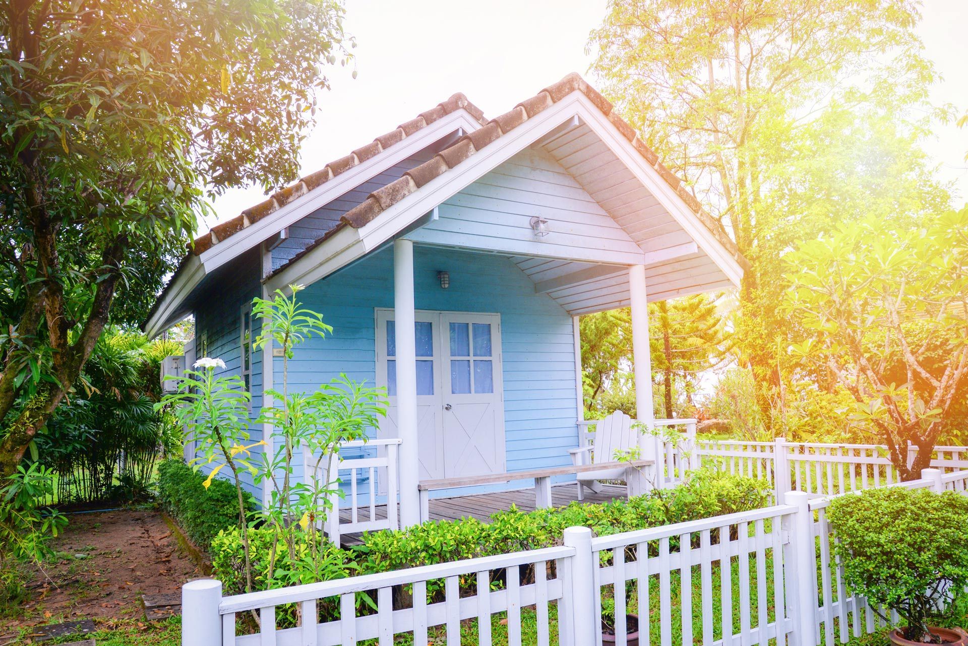 Small blue cottage with white picket fence, surrounded by lush greenery and warm sunlight.