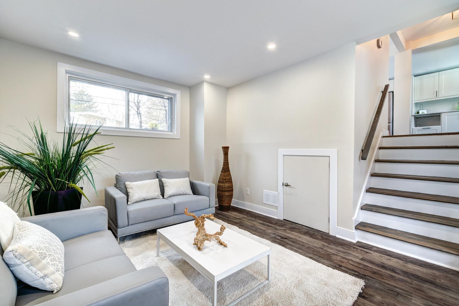 Bright living room with gray sofas, white coffee table, rug, plant, and staircase to a mirror wall.