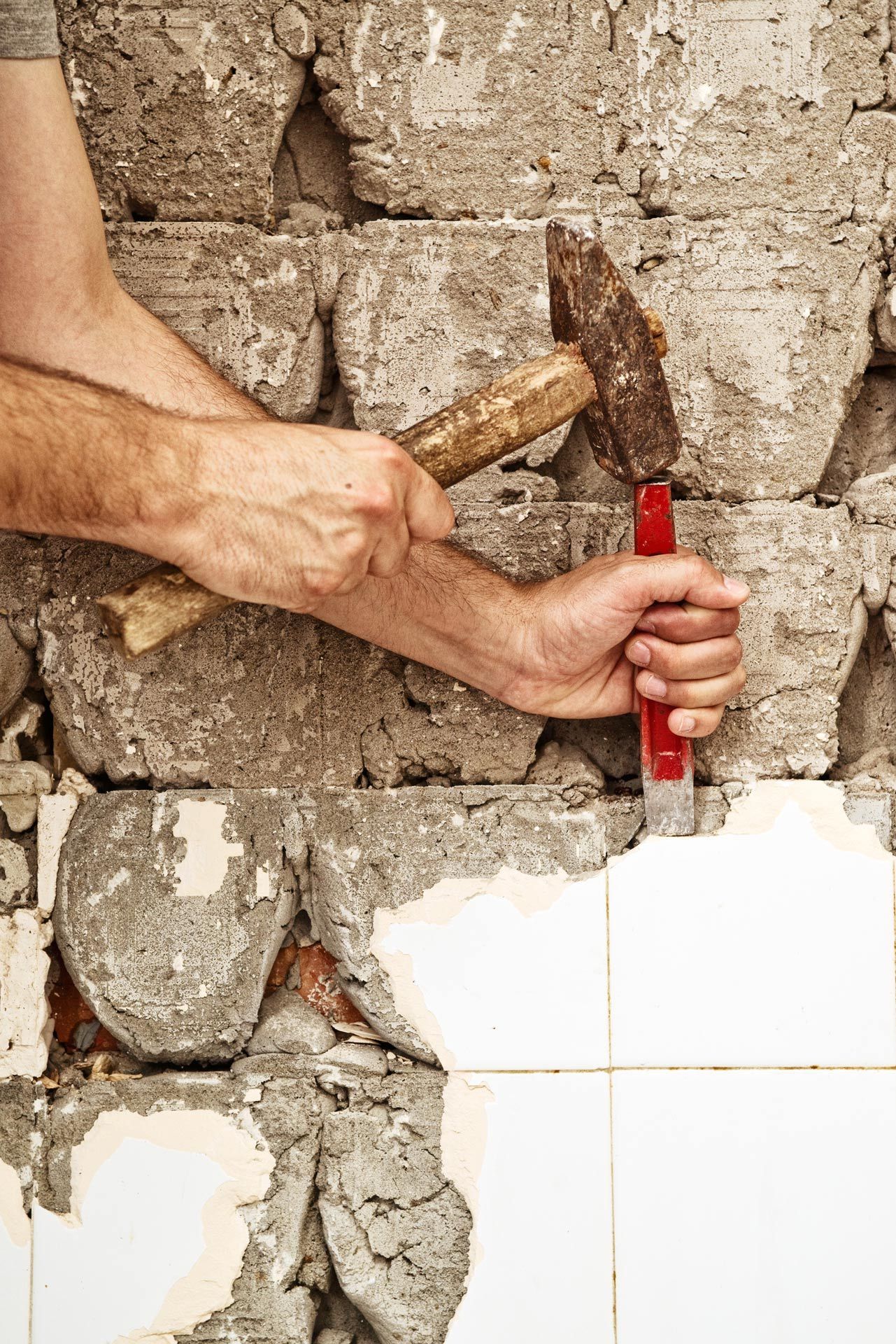 Hands with a hammer and chisel removing plaster from a brick wall beside white tiles.