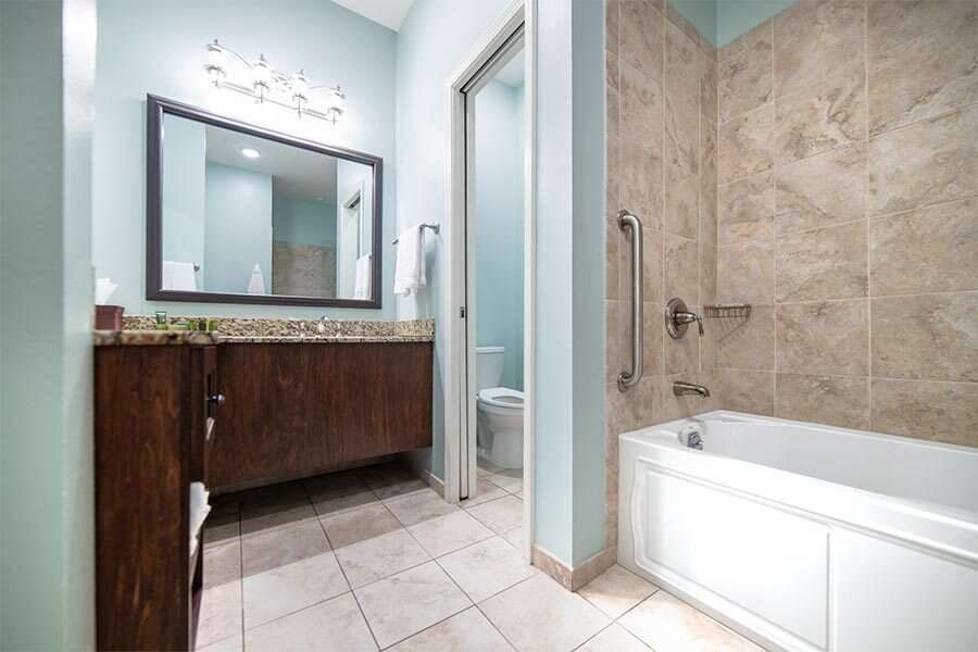 Bathroom with vanity, tub, and toilet. Light blue walls, brown vanity and tile. White tub.