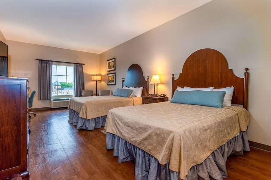 Two beds in a hotel room; brown headboards, beige bedding, wood floor, window with gray curtains.