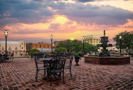 Sunset over a brick patio with wrought iron tables and fountain, buildings in the distance.