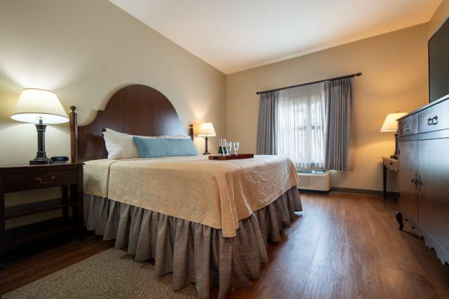 Hotel bedroom with queen bed, nightstands, and dresser, hardwood floors, neutral tones.