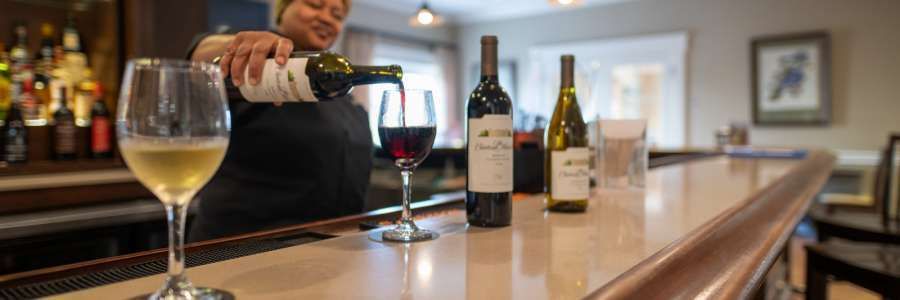 A bartender pours red wine into a glass; bottles of wine sit on the bar.