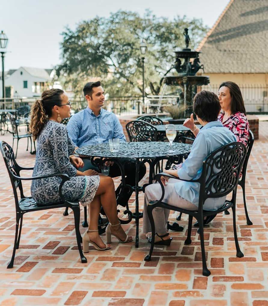 Four people seated around a table on a patio, conversing. Fountain in background.