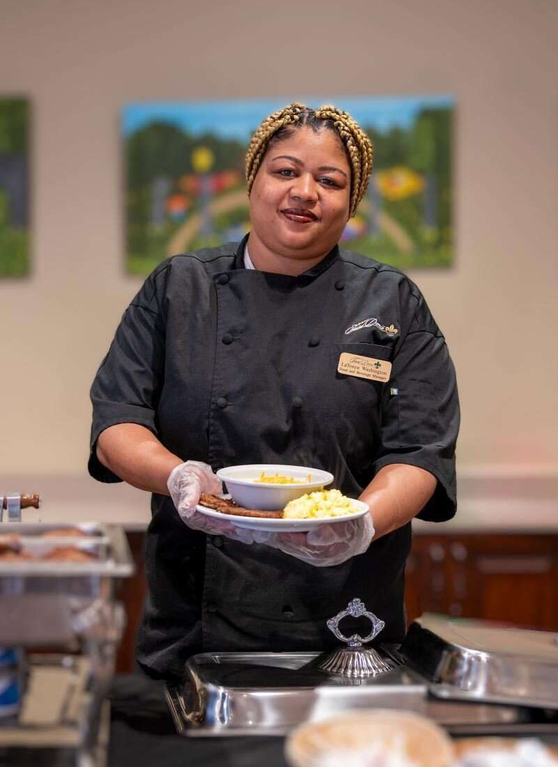 Chef in black uniform holds a plate of food in front of a buffet.