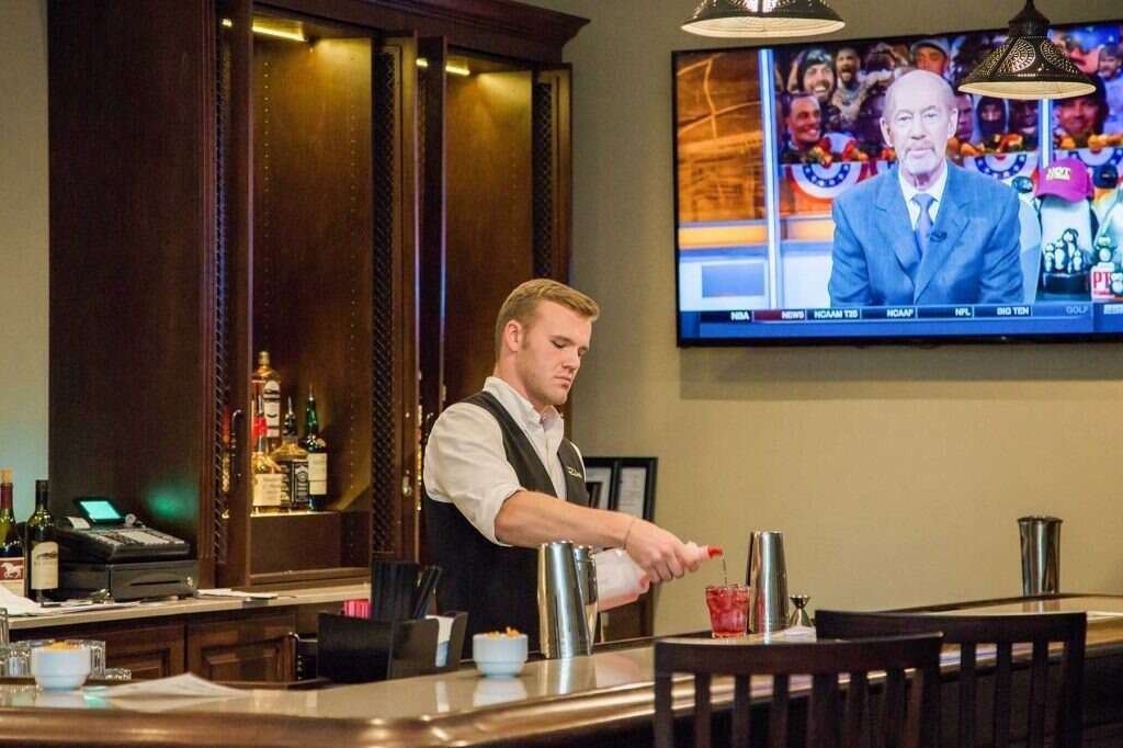 Bartender pouring from shaker, preparing a drink at a bar with a TV screen showing a sports broadcast.