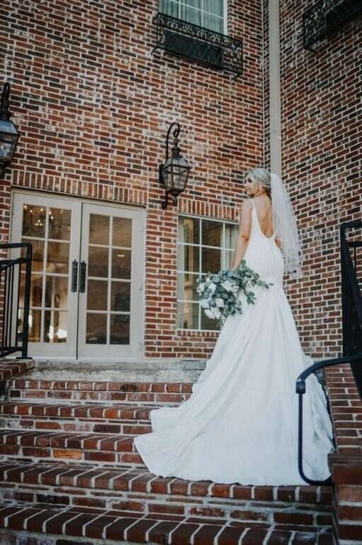 Bride in white dress and veil, holding flowers, poses on brick steps in front of a building.