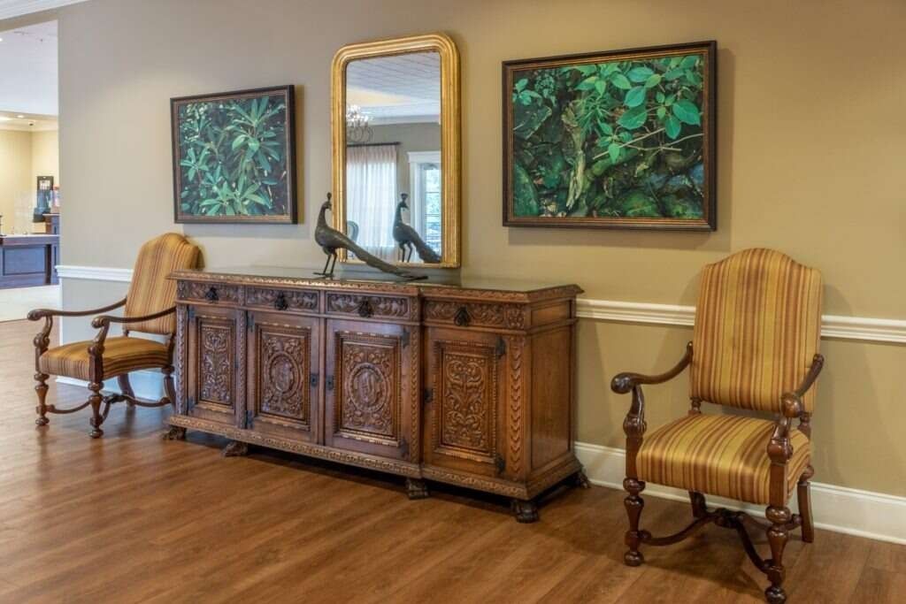 Ornate wooden cabinet with art, mirror, and chairs in a warm-toned room.
