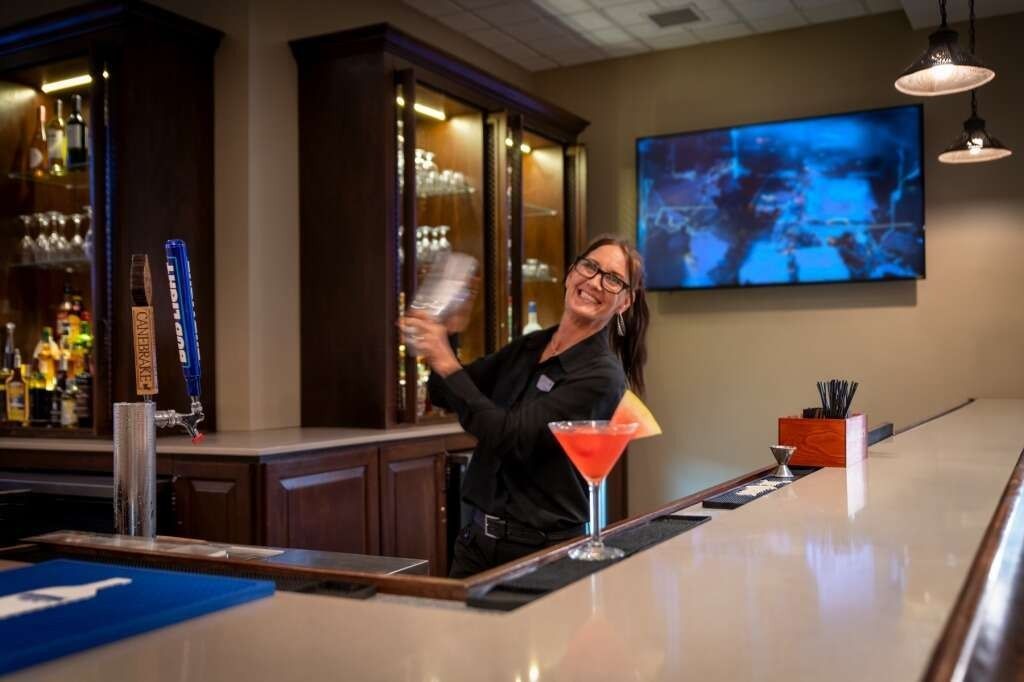 Bartender smiling, mixing cocktail behind bar. Dark wood cabinets, TV, glasses, tap handles visible.