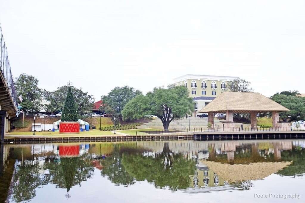 Park with reflections on water. Trees, pavilion, and buildings reflected. Overcast sky.