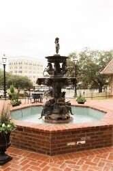 Fountain with tiered levels, bronze figures, in a brick-lined pool on a patio, with buildings in the background.
