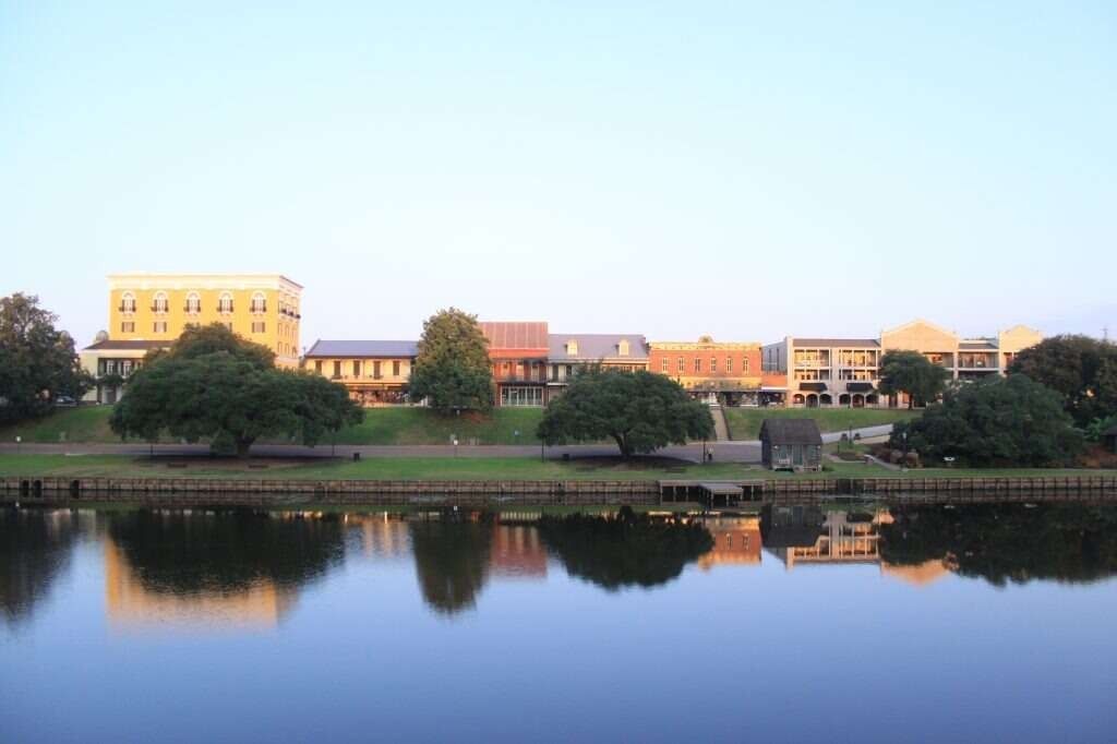 Waterfront view of buildings reflected in calm water; trees line the shore under a clear sky.