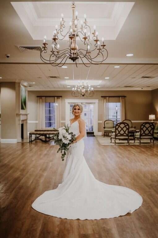 Bride in a white gown holding bouquet, standing in a ballroom with a large chandelier.