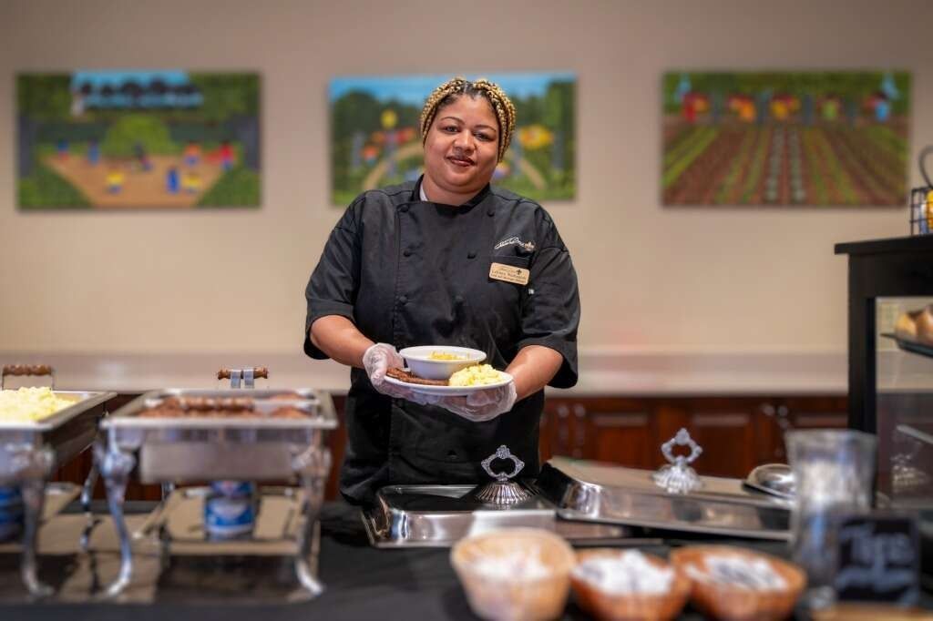 Woman in chef's uniform serving food at a buffet, smiling. Paintings and serving dishes in the background.