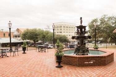 Brick plaza with a fountain, tables, chairs, and street lamps. Buildings and trees in the background.
