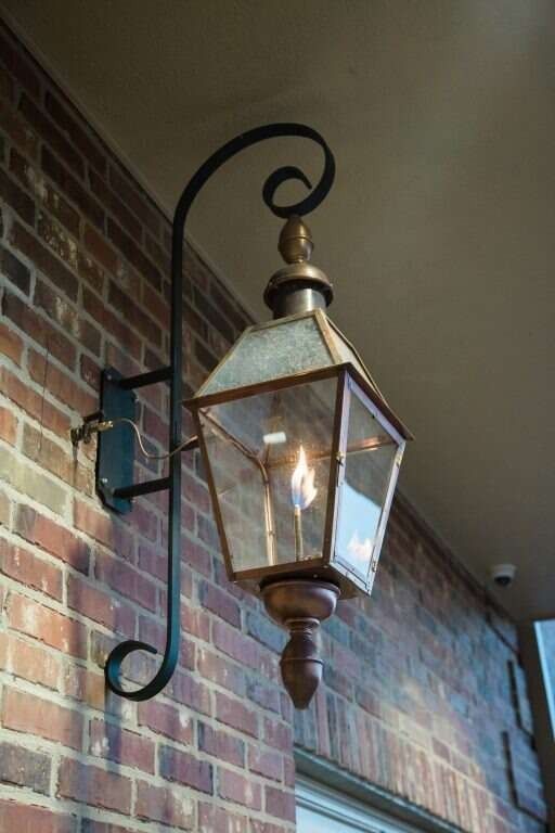 A copper and glass gas lantern with an ornate black bracket mounted on a brick wall.