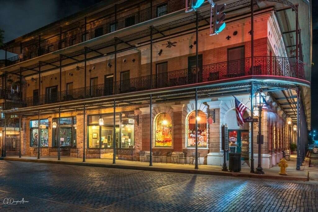 Nighttime view of a historic building with wrought-iron balconies and illuminated storefronts on a cobblestone street.