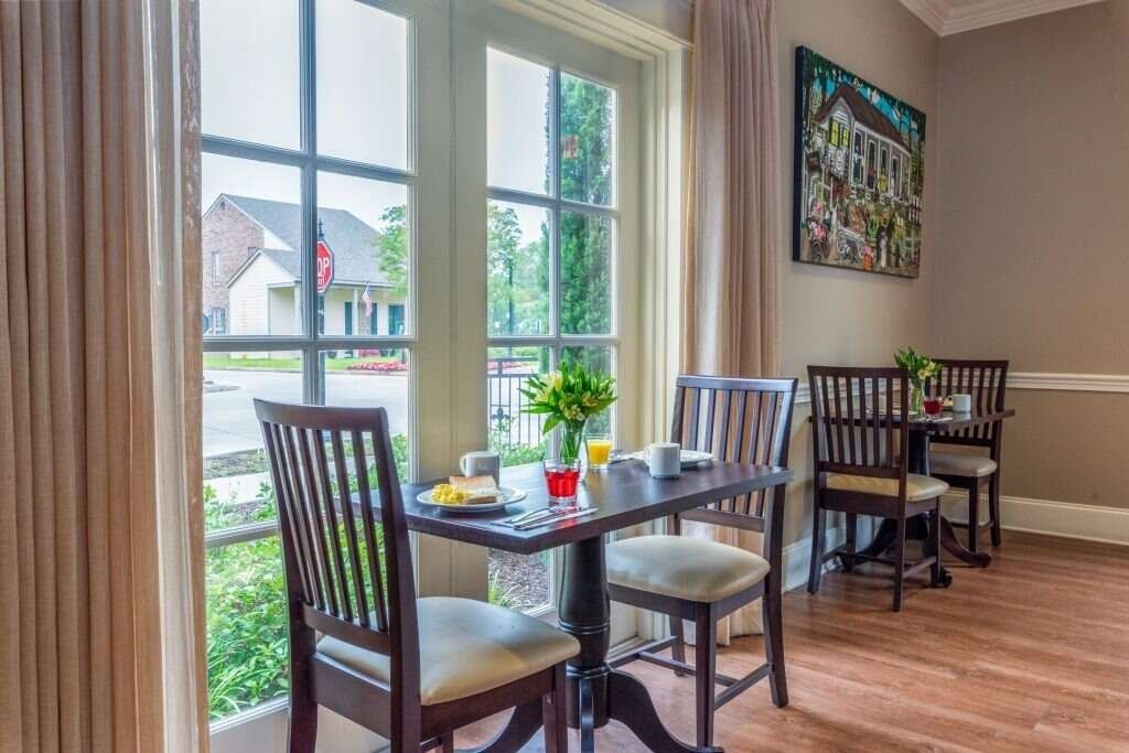Breakfast table by a window with two chairs, a view of the street and houses, and a colorful artwork on the wall.