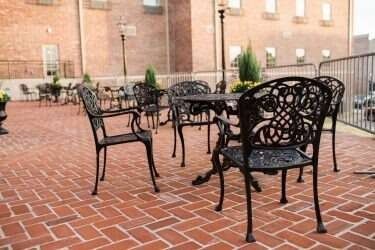 Outdoor seating area with ornate black metal furniture on a red brick patio; a building in the background.