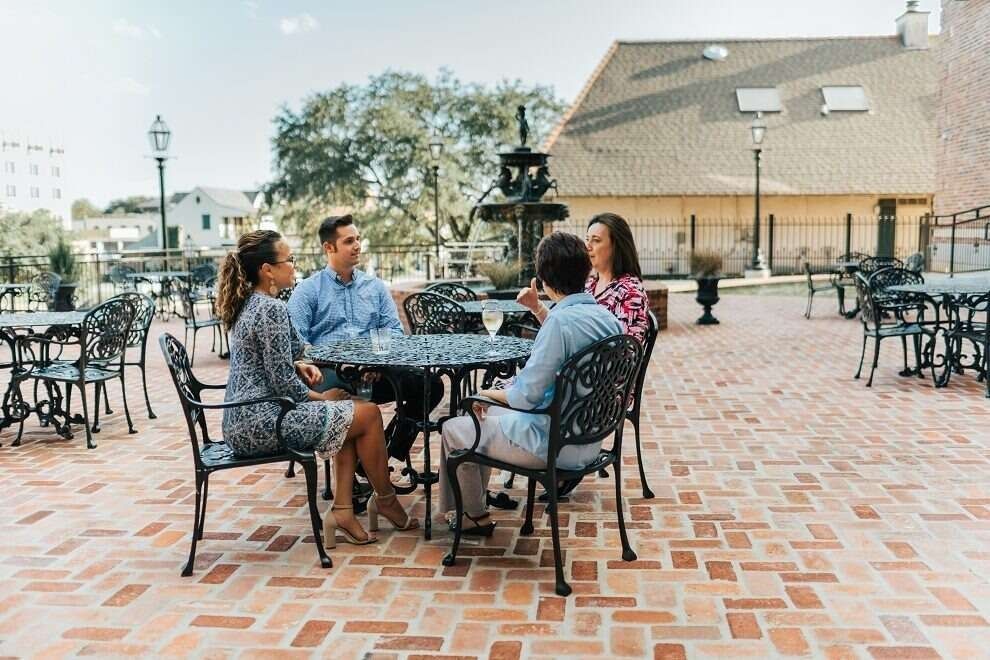 Four people seated at an outdoor table, brick patio, talking; fountain and buildings in background.