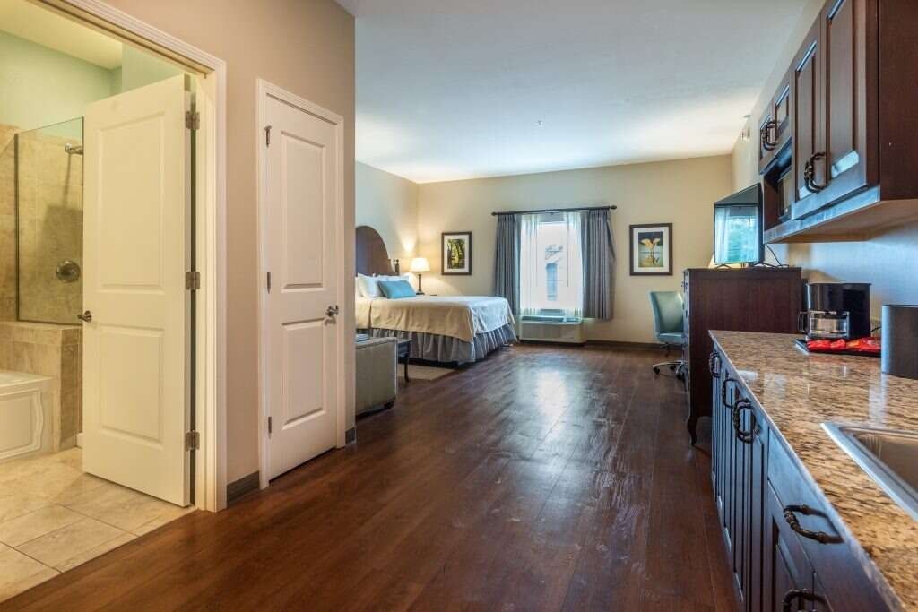 Studio apartment interior with a bed, kitchen, and bathroom. Dark wood floors, neutral walls.