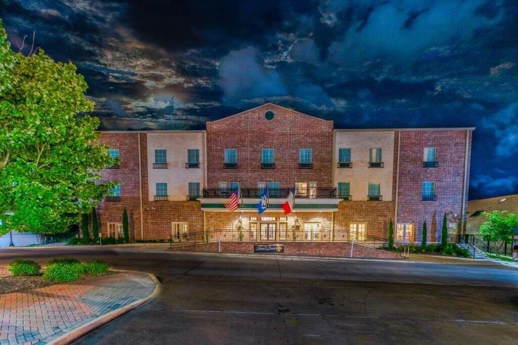 Hotel exterior at dusk with lights on, red brick facade, flags, and a brick paved driveway.