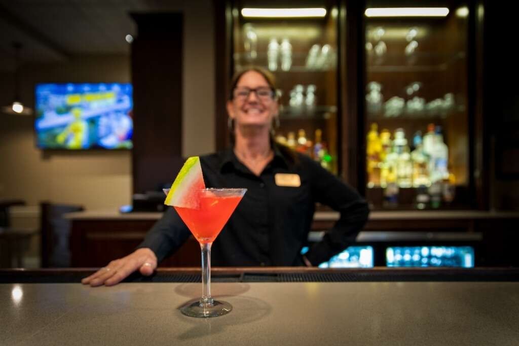 A bartender smiles, standing behind a bar with a watermelon cocktail; bar with liquor and TV in the background.