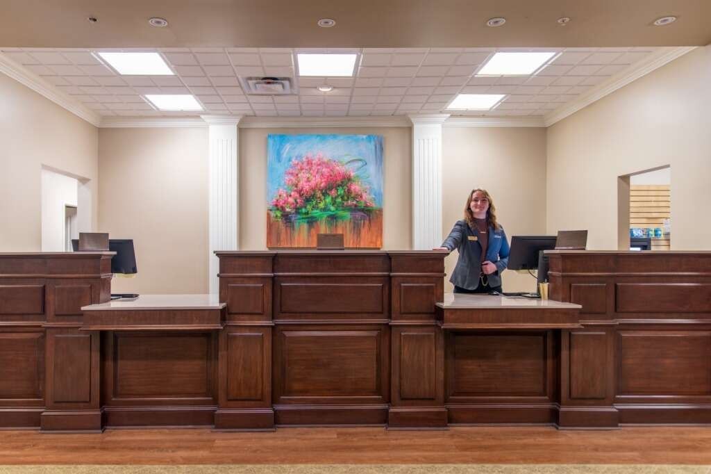 Woman at wooden reception desk in a well-lit office; painting of flowers behind her.