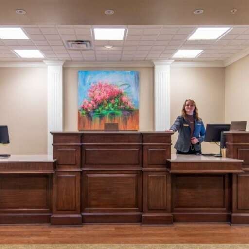 Receptionist at wooden desk, painting of pink flowers above. Beige walls, columns, and bright ceiling lights.