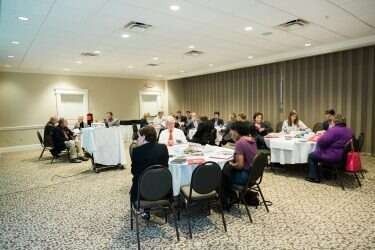 People seated around tables in a conference room, engaged in a meeting.