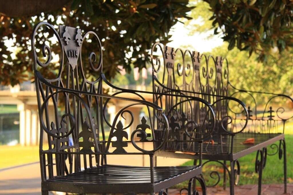 Ornate black metal bench under a tree, by a path, with a bridge in the distance.