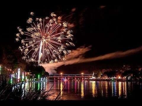 Fireworks display over a body of water, reflecting colorful lights and a bridge in a dark night.
