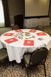 Round table set for a meeting with red folders, chairs, and a projector screen in a hotel room.