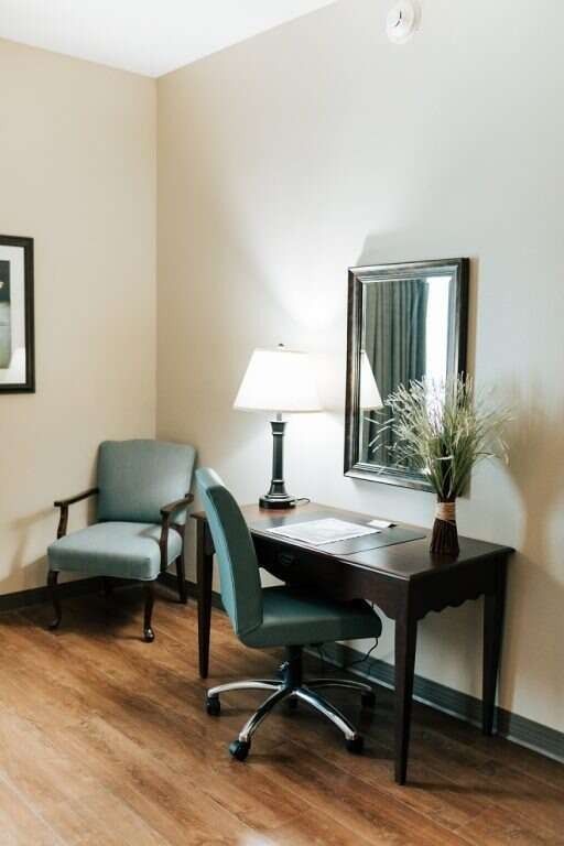 Desk with chair, lamp, and mirror in a room with a chair in the corner, wood floors, beige walls.