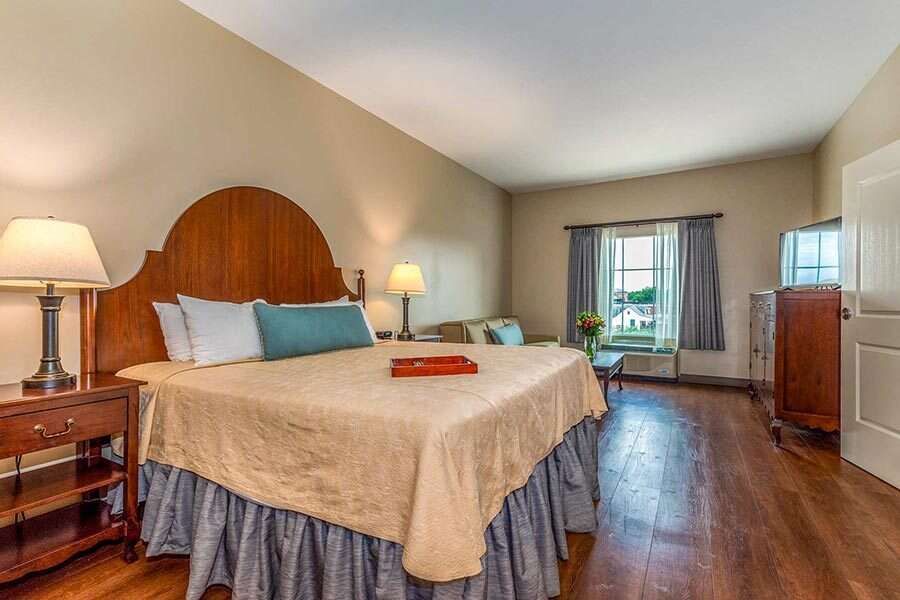 Hotel room with a large bed, nightstand, sofa, and window with curtains. Wooden floors and beige walls.