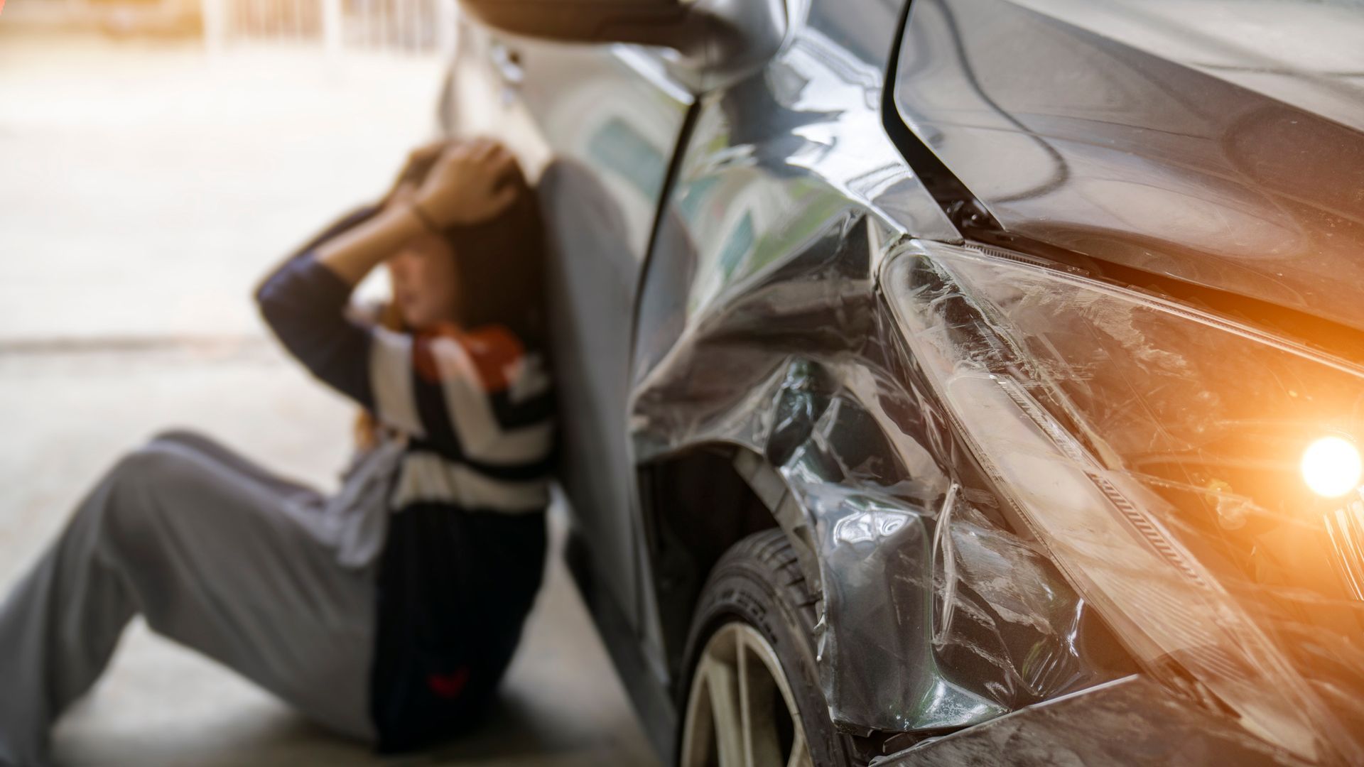 A woman with hands on her head sits by a car with a crumpled front-right fender 